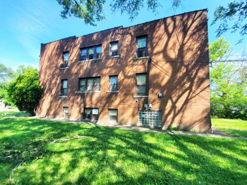 The sun-dappled side of a two-story-plus-basement brick multifamily apartment with a green lawn.