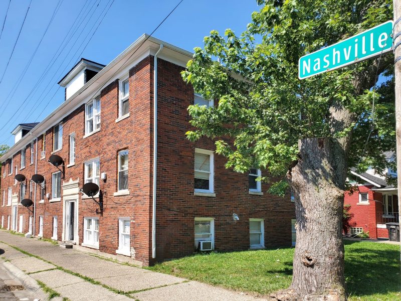 A corner view of a 3-story brick 9-unit apartment buiding with white-trimmed windows and a deciduous tree and green lawn on the side. A street sign saying "Nashville" is visible in the top right corner of the image.