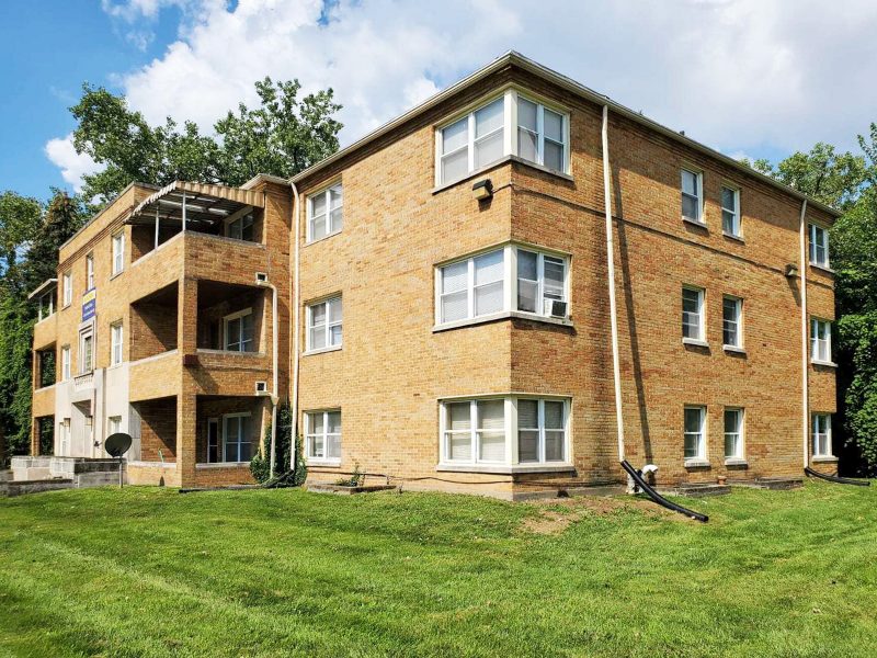 A corner view of a 12-unit multifamily apartment done in tan brick, with a large green lawn in front.