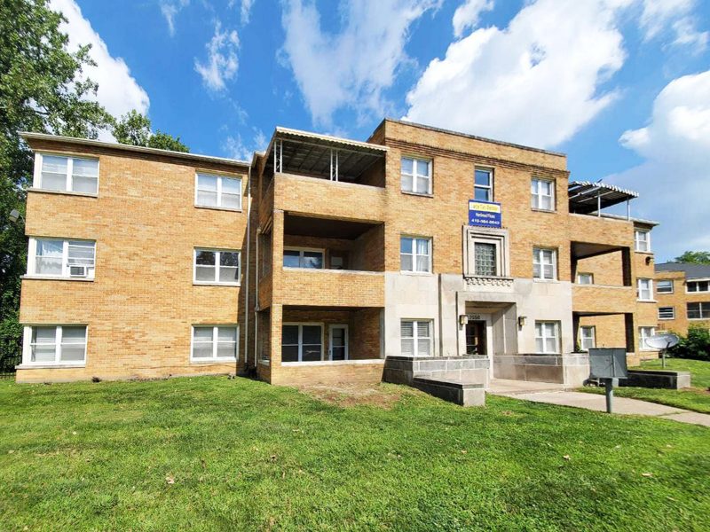 The facade of a 12-unit multifamily apartment done in tan brick, with a large green lawn in front.
