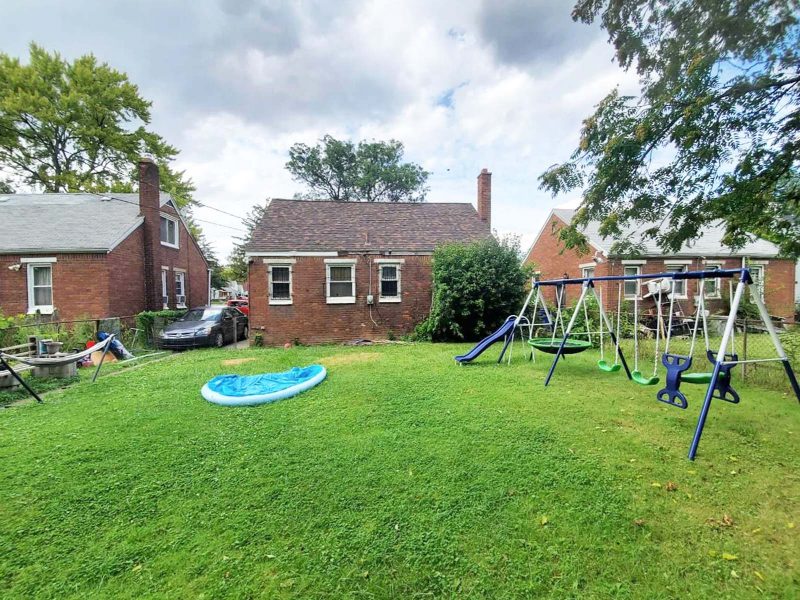 A wide view of the large green lawn behind a brick single-family home, including an empty inflatable pool and a large swingset.