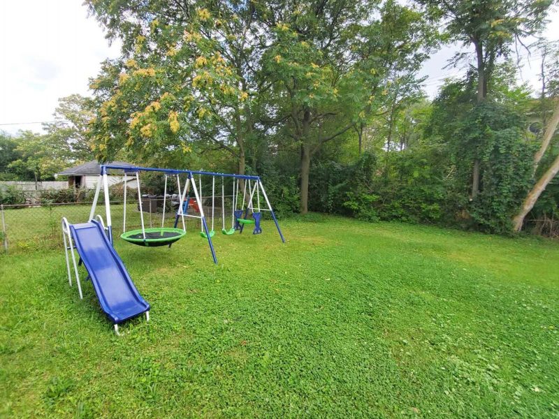 A view of a green lawn with a large swingset on the left side, shaded by deciduous trees.
